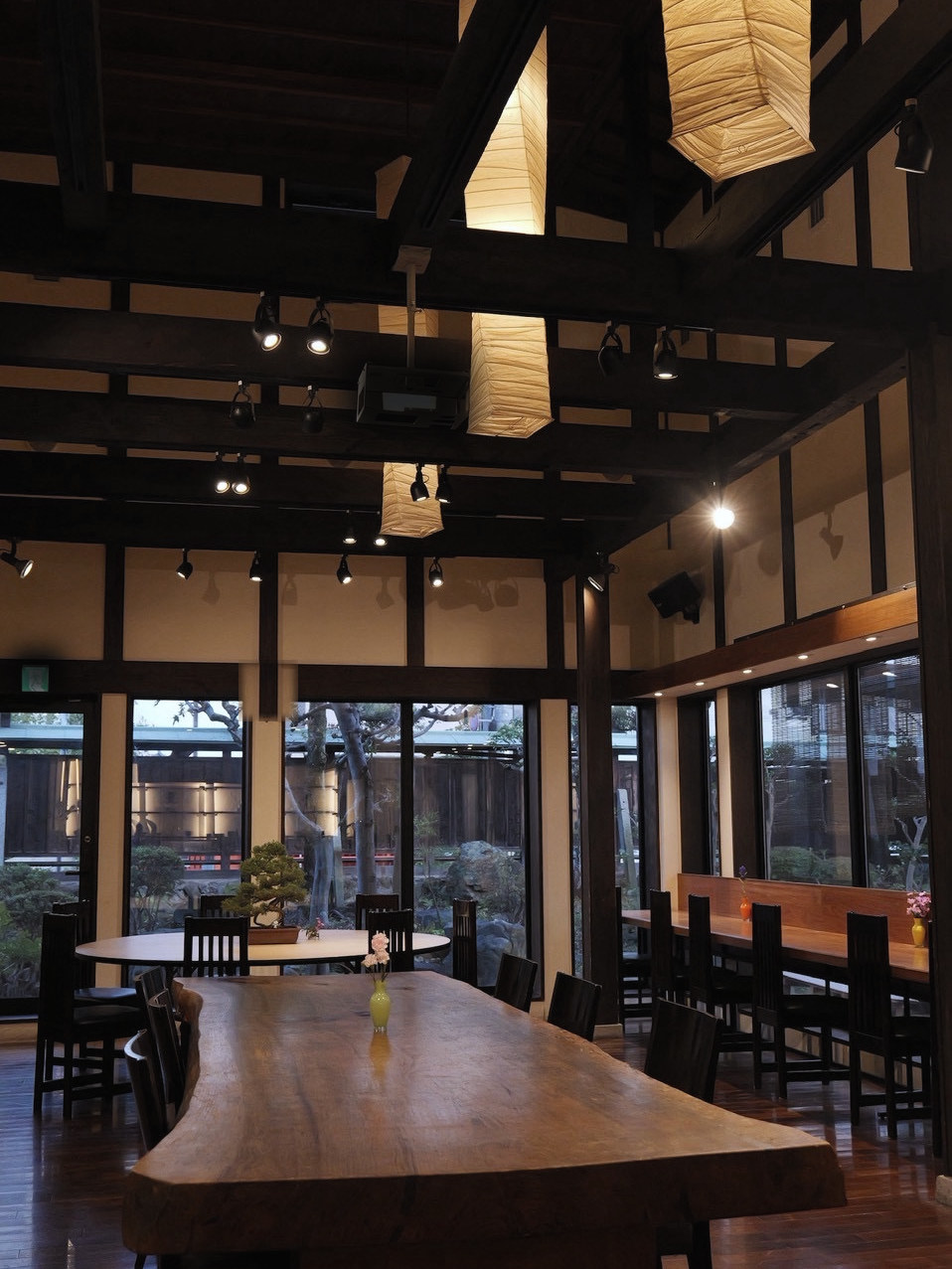 和紙の吊り照明が灯る夕暮れの店内。黒い梁の下に、一枚板のテーブル